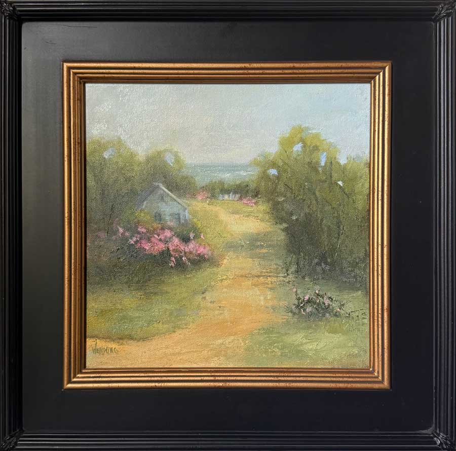 A framed landscape painting featuring a dirt path leading through green trees to a small white house with pink flowers, set in a black frame with a gold inner border.