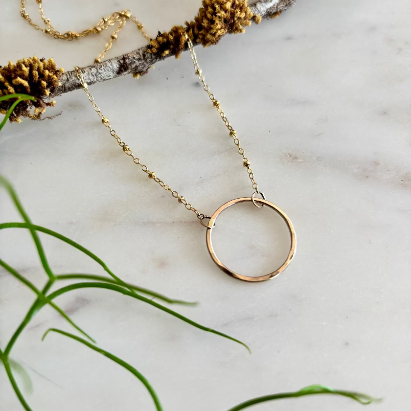 Gold necklace with a circular pendant on a marble surface with greenery