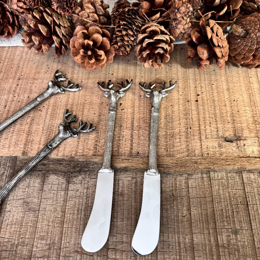 Three silver deer-head spoons on a wooden surface with pine cones.
