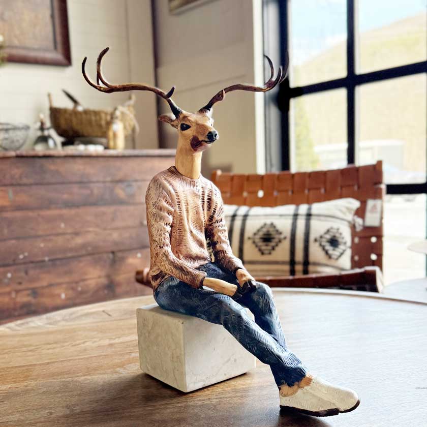 Deer figure wearing a sweater and jeans sitting on a block in a room with wooden floor and furniture.