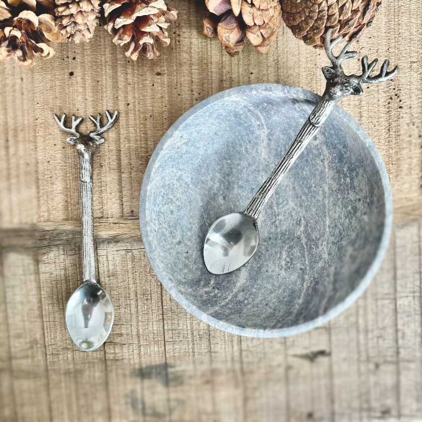 Two silver spoons with deer heads on a wooden surface with pine cones.