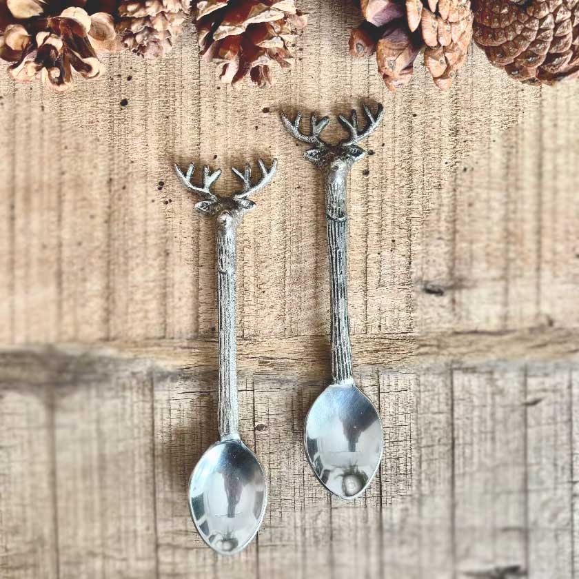 Two silver spoons with deer head designs on a wooden surface with pinecones.