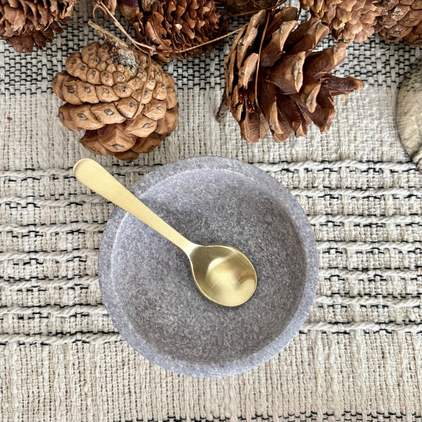 Gray stone bowl with a gold spoon on a textured surface with pine cones.
