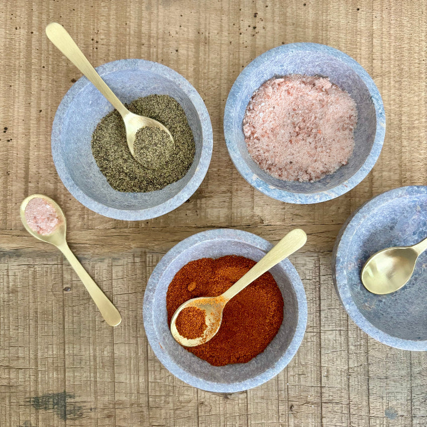 Four bowls with different spices and gold spoons on a wooden surface