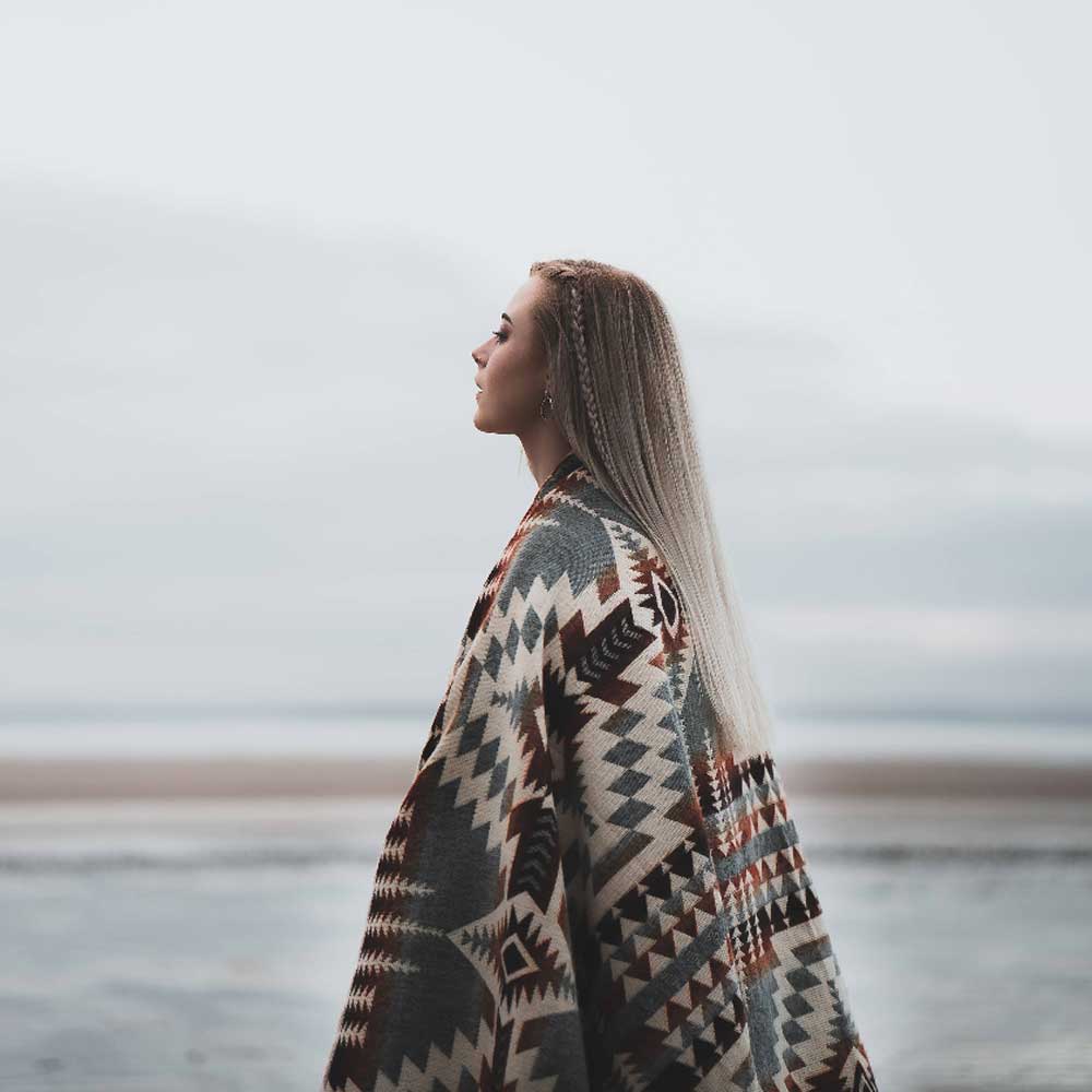 Person wearing a patterned blanket standing on a beach with a cloudy sky.