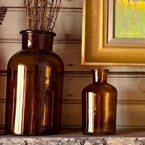 Two brown glass bottles on a wooden surface with a framed picture in the background.