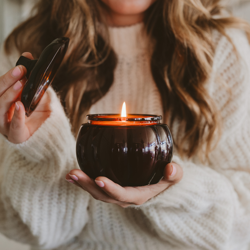 Person holding a lit candle in a dark glass holder with a blurred background