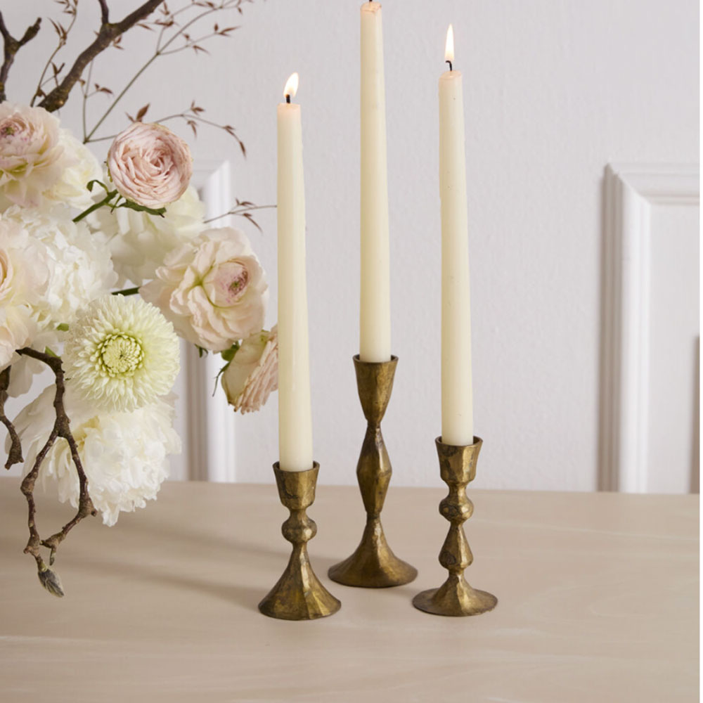Three tall candles in decorative brass candlesticks with a floral arrangement on a light surface.