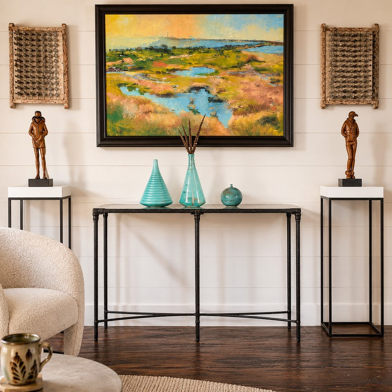 A framed landscape painting hangs on a white shiplap wall above a console table in a styled living room. The painting depicts a marshland scene with blue waterways and warm golden-green grasses.