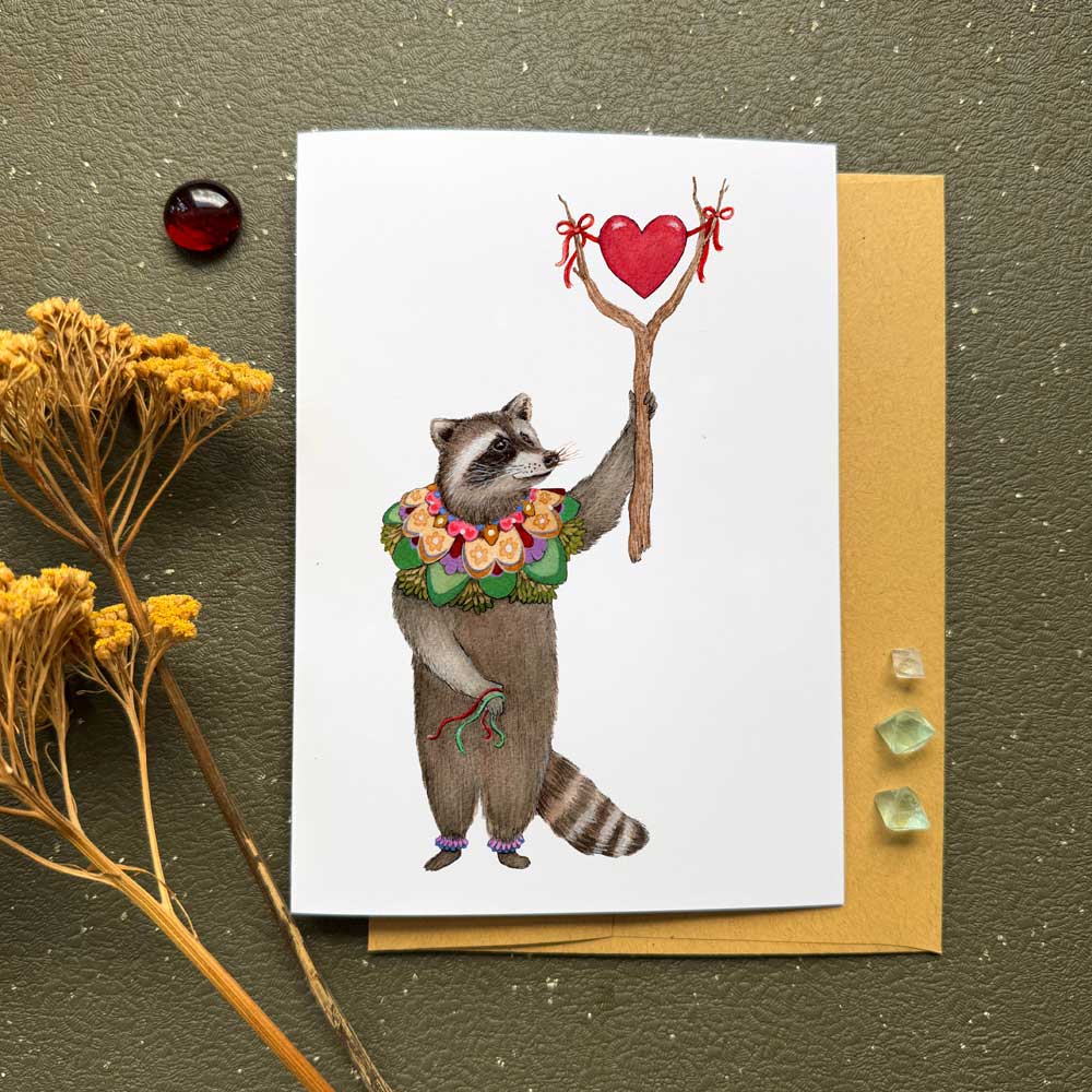 Greeting card with a raccoon holding a heart and flowers on a textured surface.
