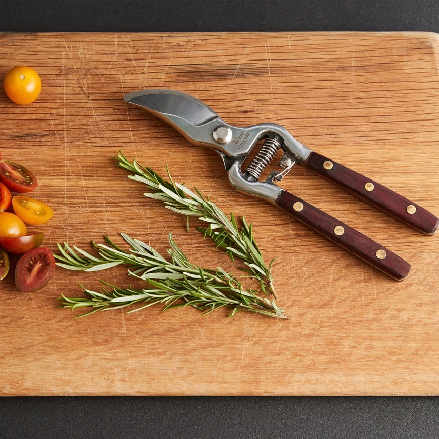 A pair of pruning shears with dark brown wooden handles and silver metal blades, laid diagonally on a wooden cutting board next to fresh rosemary sprigs and halved cherry tomatoes.